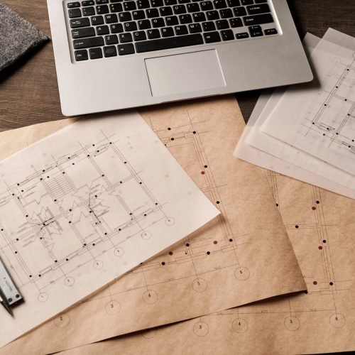 Laptop, handtool, pen and cordless earphones surrounded by unfolded blueprints with sketches of new building drawn by modern engineer